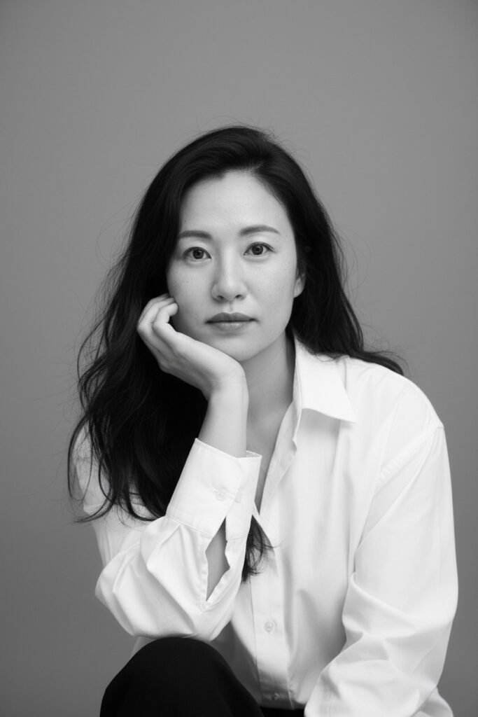 Black-and-white portrait photo of a person with long hair wearing a white shirt, resting their chin on their hand against a plain background.